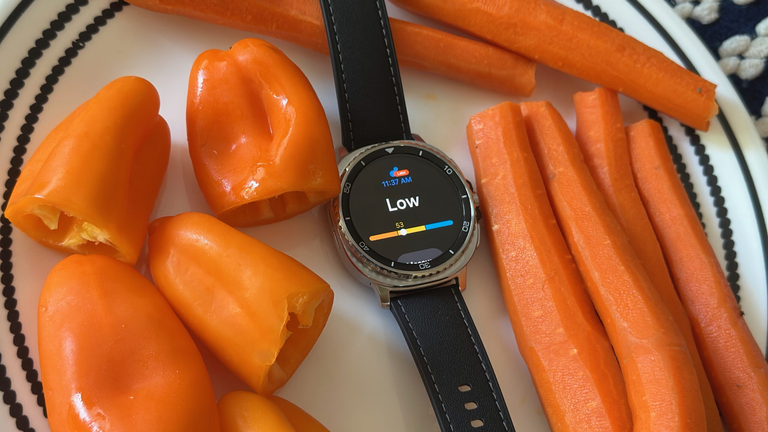 The Samsung Galaxy Watch 8 Classic sitting on a plate surrounded by bell peppers and carrots, the display showing the Antioxidant widget with a &quot;Low&quot; score.